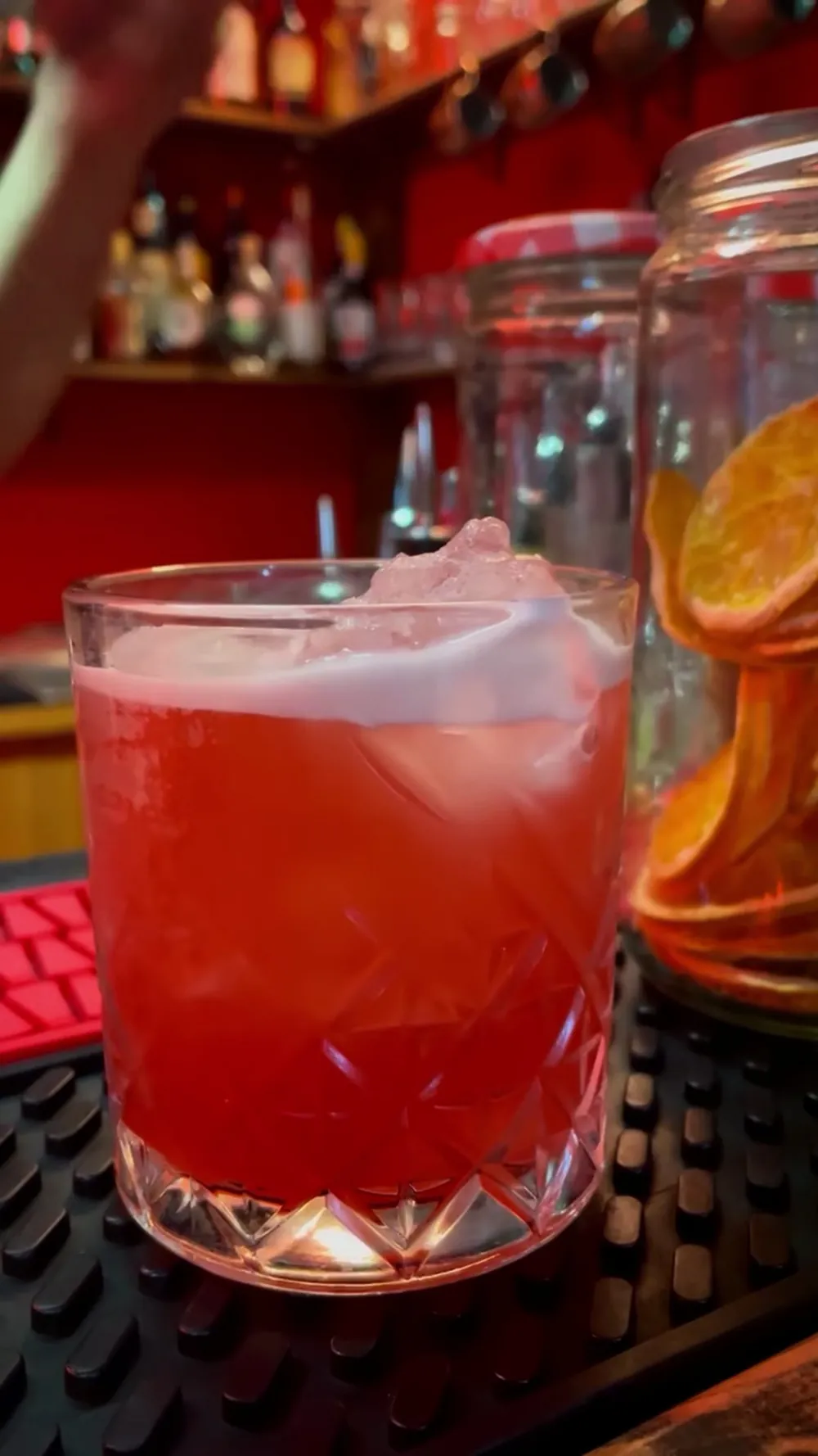 Dietro il banco: un cocktail rosso con ghiaccio, i barattoli di vetro con le arance essiccate e la fila delle bottiglie