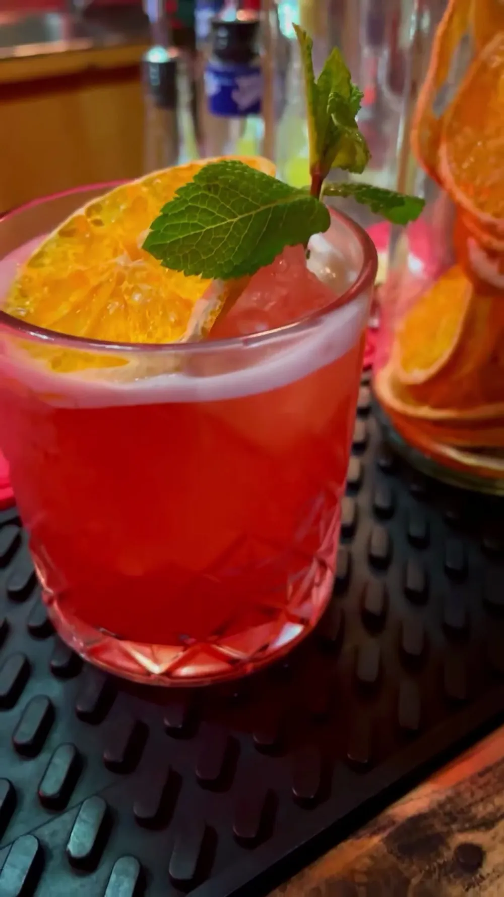 Il bancone del Moonshine sotto la luce rossa: un cocktail rosso con fetta d'arancia e menta, accanto a un barattolo di vetro pieno di arance essiccate