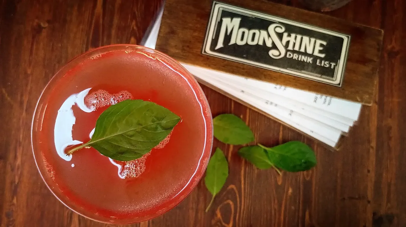Cocktail rosso visto dall'alto con una foglia di basilico a galla, accanto alla drink list