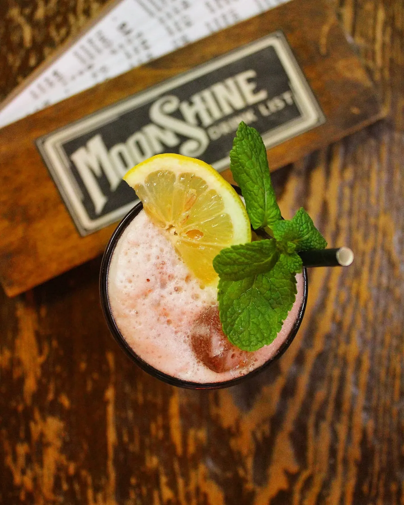 Cocktail rosa con fetta di limone e menta visto dall'alto, accanto alla tavoletta della drink list