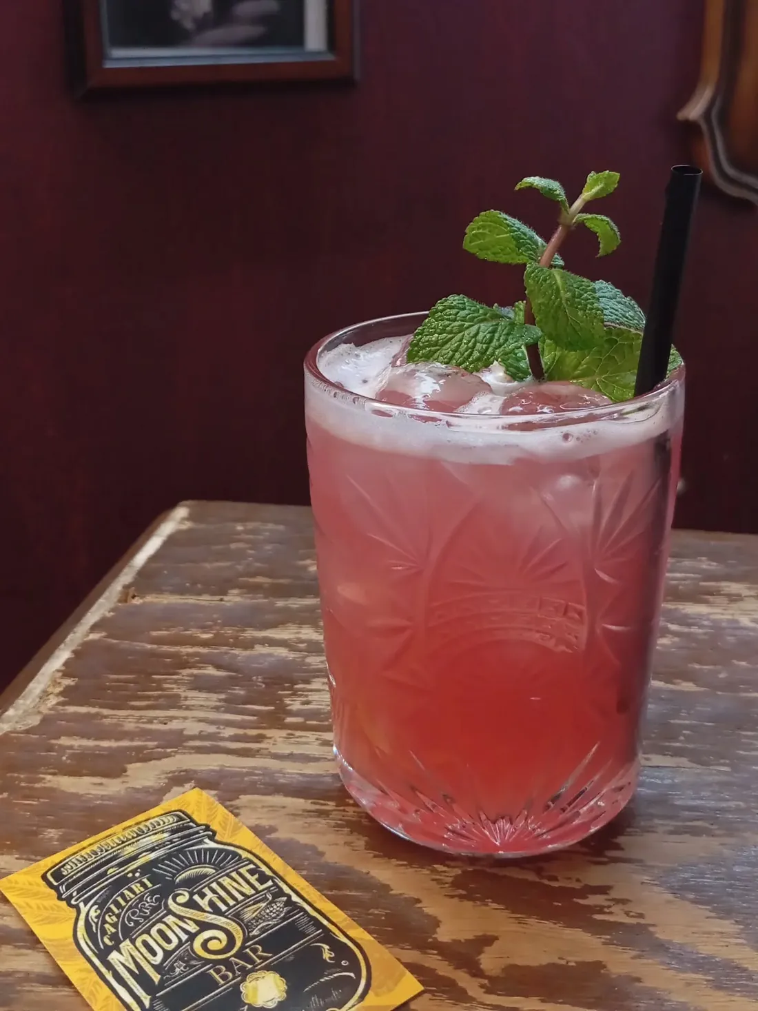 Cocktail rosa con ciuffo di menta in bicchiere di cristallo molato, accanto al biglietto giallo del Moonshine