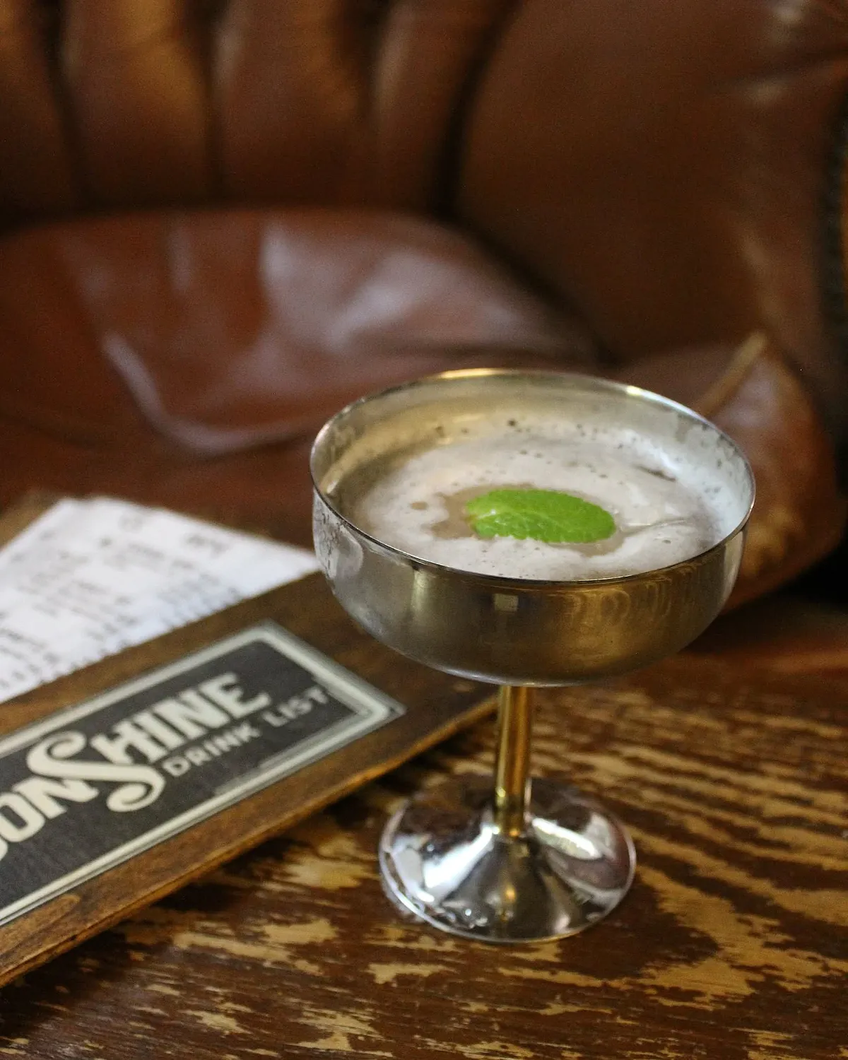 Coppa in metallo con un cocktail schiumoso e una foglia di menta, davanti a una poltrona in pelle marrone