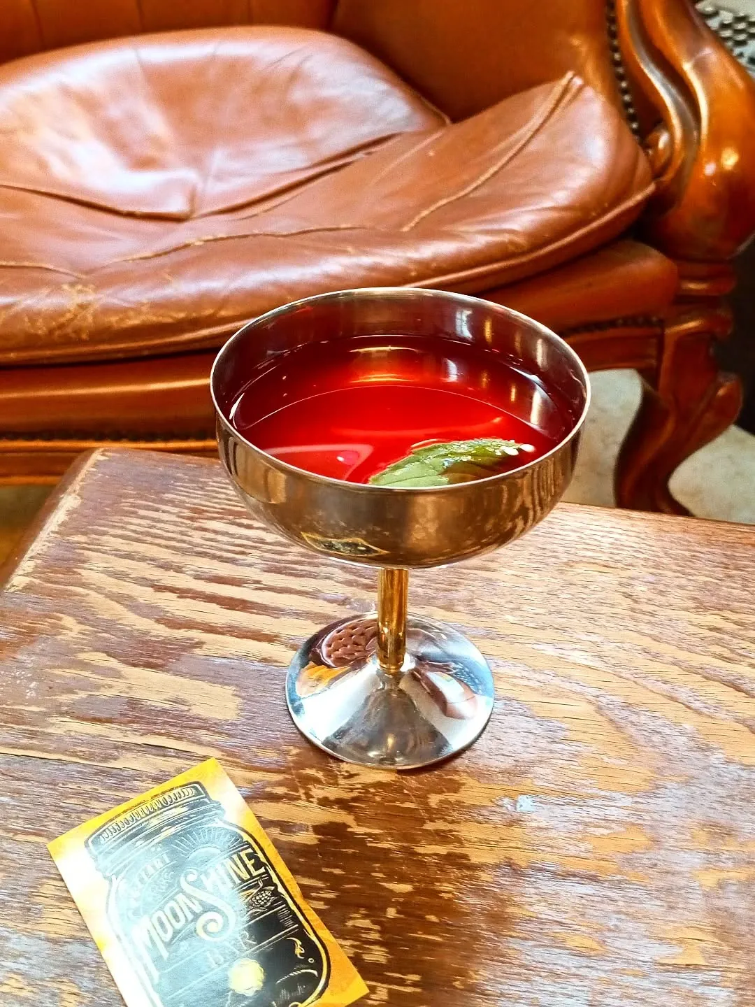Coppa in metallo con un cocktail rosso rubino, su tavolino di legno davanti a una poltrona in pelle