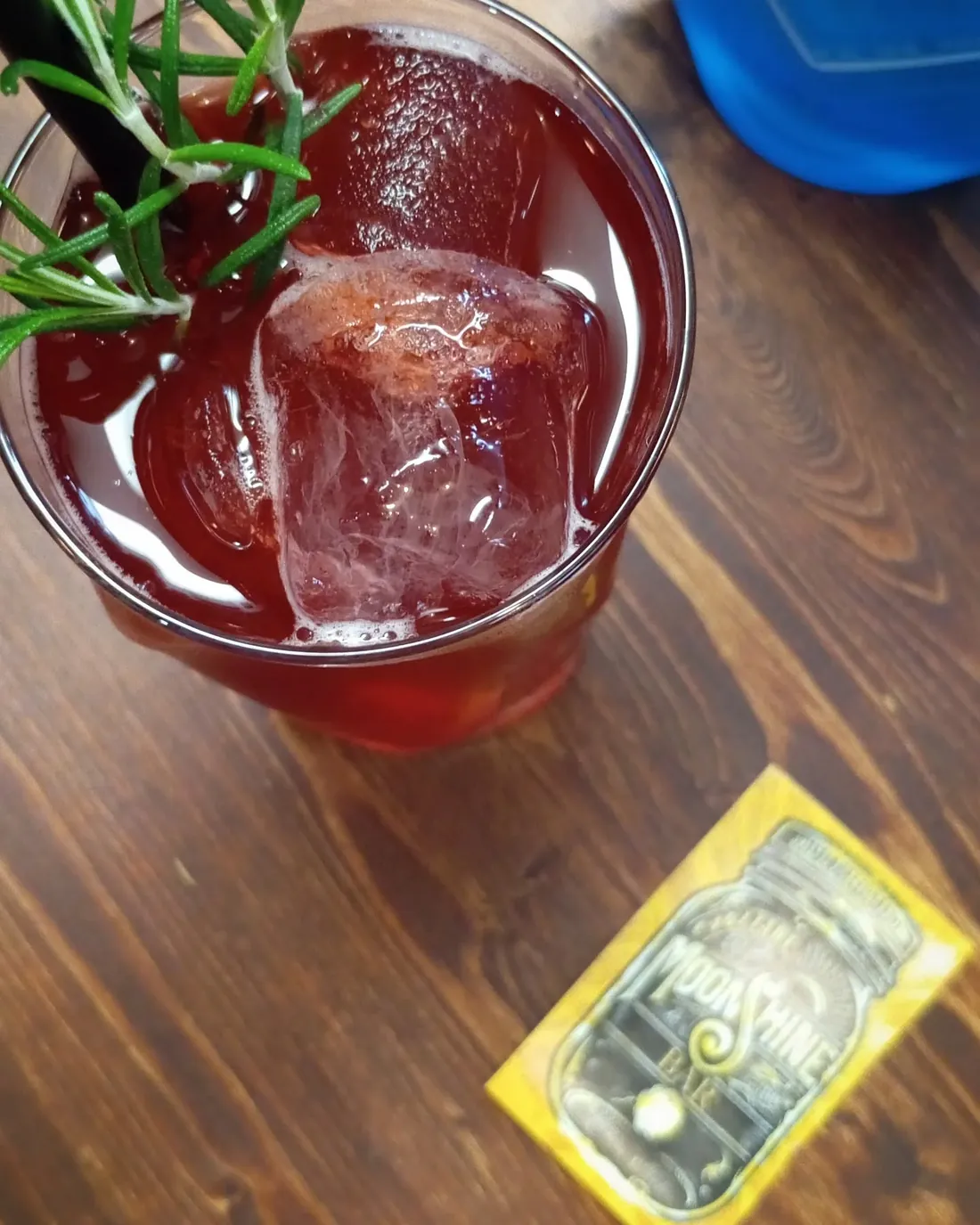 Cocktail rosso scuro con ghiaccio grande e rosmarino, accanto al biglietto giallo del Moonshine