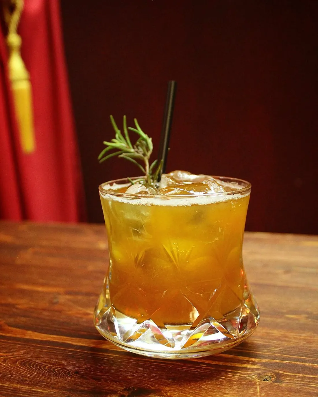Cocktail ambrato in bicchiere di cristallo molato con rametto di rosmarino, su tavolo di legno scuro