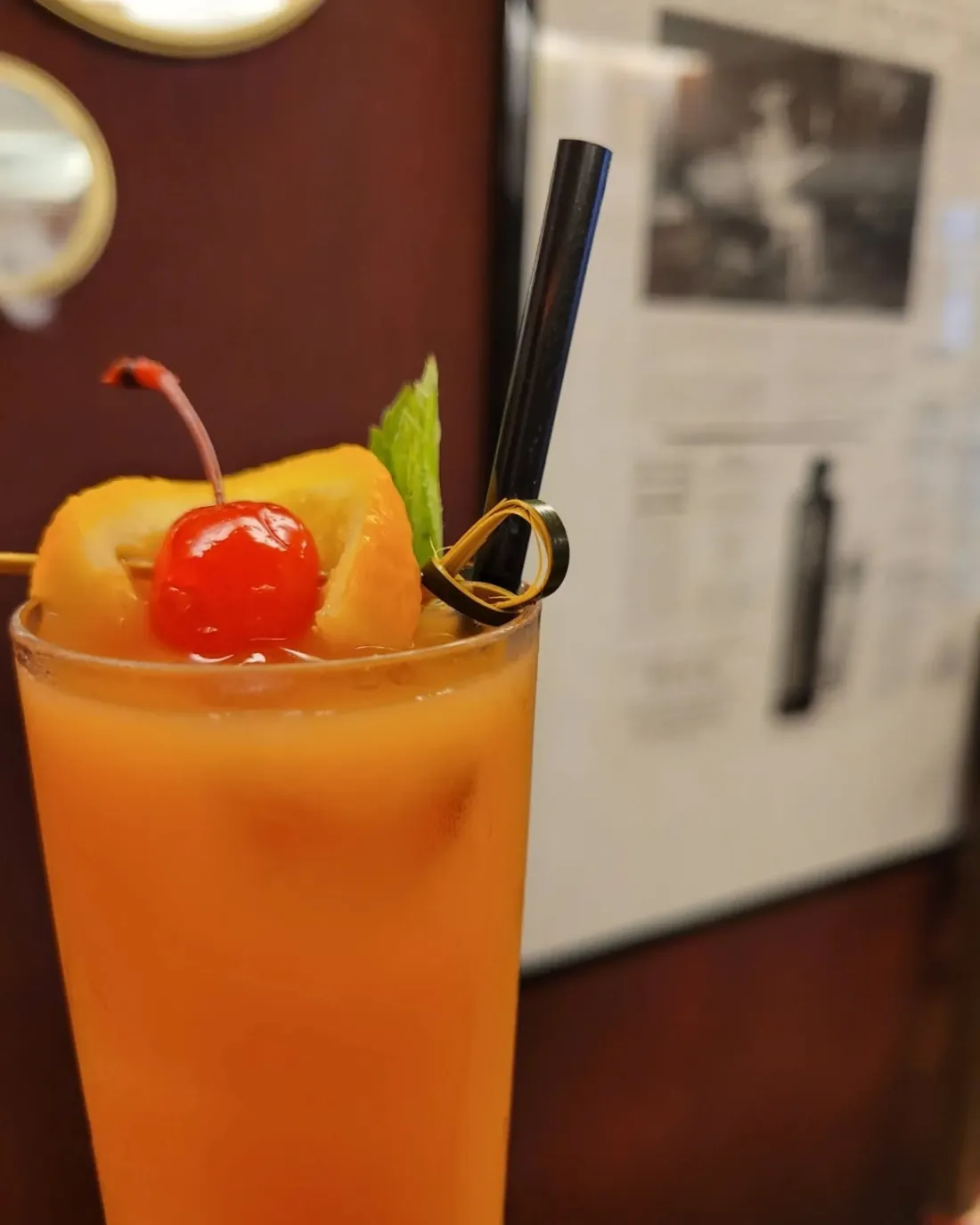 Long drink arancione con ciliegia e cannuccia, davanti a una cornice appesa al muro